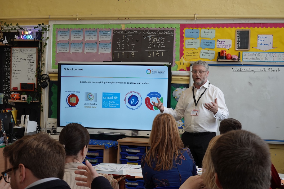 Mr Anderson at the front of a classroom, describing the Shaftesbury Park Primary School approach to international visitors